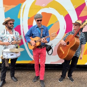 Photo of three performers standing infront of an abstract colourful mural, the performers are wearing colourful clothing and hold a drum, guitar and large guitar