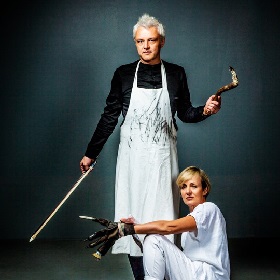 Photo of Jody Graham crouching with a makeshift arts multi tool artists glove. it looks like a black glove with different implements at the end like a brush and other mark making items. She wears all white and has a short blond bob, Mark Cauvin stands behind her holding a violin bow , he has a white apron with black marks on it and holds a stick in his other hand