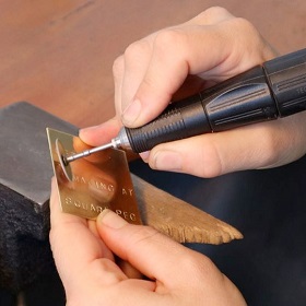 Photo of two white hands engraving a gold plate with an electrical engravers tool