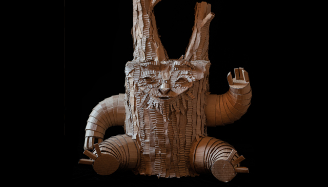 Photo of a cardboard sculpture of a fictional tree stump man, very cute, the stump has cylindrical consertina type legs, similar arms and two stalks coming out of its head, the stump has a smiling face all made from cardboard
