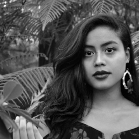 Black and white photo of person with long dark hair, large hoop earrings, the background is full of palm tree leaves 