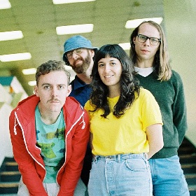 Photo of the musical group the Beths, four people are in the shot from head to thighs, one is at the front in a yellow top tucked into light denim jeans, to their left is another in a red hoody with a moustache and bent over pose, in the background another wears a blue denim fishermans hat, glasses and a beard, next to them is the last person who appears tall and slender and wear black framed glasess, long hair and a  green long sleeved v-neck jumper whit a white tshirt underneath - the photo has a playful feel