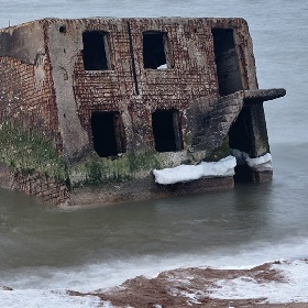 Photo of a Brick two story small building that looks very dilapidated - the structure of the building and the raw concrete are all that are left - the building is sinking into the water and has moss or seaweed that has grown at the bottom where its been close to the water, it has some stairs that are in a door opening from the top level and run on the outside below a window, there are two window holes on the top and bottom level is mirrored below. The water is a murky brown and there are some brown rocks that the waves have crashed onto