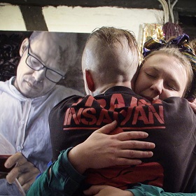 Promotional movie image shows photo of woman hugging man (his face is facing other way) in the background is a painting of the same man in black and white