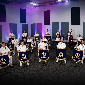 Photo of the Royal Australian Navy Big Band wearing white uniforms sitting ijn front of music stands with official gold fringed and navy coloured music stands, the word Australia is written below a navy looking emblem, the lighting is purple and there are three lines of musicians with different instruments