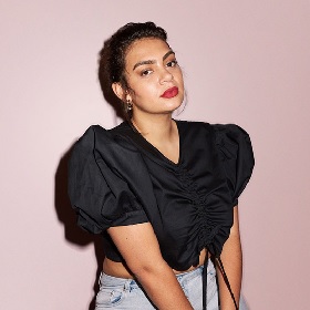 Studio photo of Thelma Plum, Thelma poses for the camera - there is a dusty pink block colour background, a black shadow outlines her left side. She wears her  dark hair loosly up and has bright red lipstick, she is beautiful and tilts her head on the side. She is wearing a puffy sleeved black top ruched at the front and pale denim jeans.
