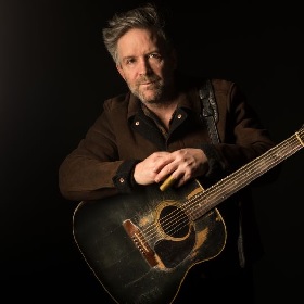 Photo of musician wearing a brown coat, they hold an acoustic guitar that looks like it has been painted black but the colour has worn off at high touch points, the musicians hands are placed above the guitar and one finger is covered in a bronze finger slide. The musician looks straight at the camera and has  grey brown short hair and facial hair