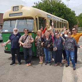 Photo of a tour group standing in front of a vintage bus, they wave and smile at the camera
