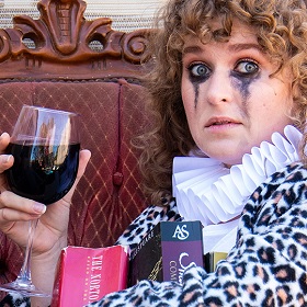Woman with curly brown hair looks at camera she has lots of black eye makeup around her eyes ) top and bottom it is also running down her face, she has a glass of red wine in her hand and books on her lap. she wear a white ruffle neckerchief and a leopard print fur top  