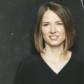 Photo (head and shoulder shot) of person with a brown bob haircut, blond highlights at front of head, broad smile, black v neck top, nose ring and black background