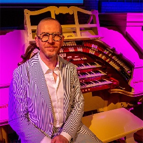 Photo of a man wearing a stiped suit and open white shirt, he wears statement tortoise shell glasses, behind him is a Wurlitzer or organ type instrument there is a purple warm glow to the photo