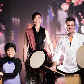 Three performers are photographed, the background shows an image of pink fruit blossoms, one oerformer sits with a kind smile on her fce, performer next to her stands with a broad happy smile, she holds a drum with red patterned sash. the third performer is a man who sits with a drumstick in one hand and a drum in another hand, he smiles and is wearing a white shirt.