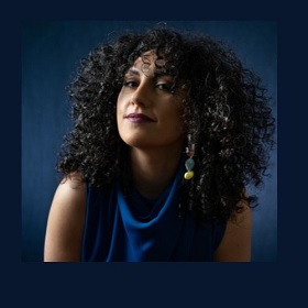 Photo of a person with olive coloured skin, they have dark curly hair that frames their face and reaches below their shoulders, they wear a blue cowlneck sleeveless top, long dangly earrings and purple-red lipstick