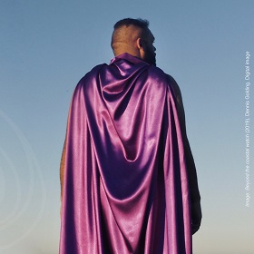 Man wears purple shiny cape with  blue sky background, he is looking into distance