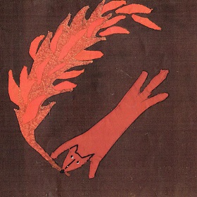 Photo of sewn red fox on brown material, fox holds a flaming red stick in its mouth, fox is stretched out and view is from above