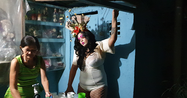 Photo of Ate Indai in performance, she wears a decorative headress made of flowers and a fan, she wears white lingerie like body suit with white satin short sleeves, she has white fishnet stockings on and looks down a table where a woman is cleaning dishes, in the background is a kitchen painted blue with crockery behind a make-shift plastic curtain