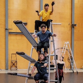 4 performance artists in workout gear balance ontop of each other with a ladder prop , one performer stands on the ground looking up at the other performers
