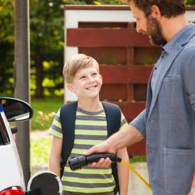 Boy in a striped green and black tshirt looks up at a man who is holding an electric vehicle charger towards an electric vehicle