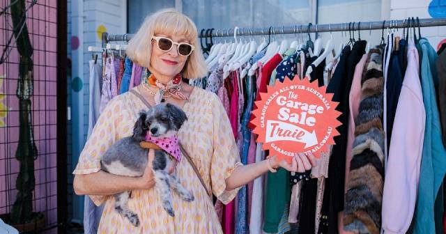 Woman with grey hair holding a small dog stands in front of a rack of clothes and holds a sign saying Garage Sale Trail