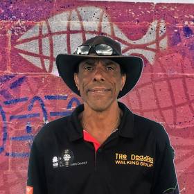 Dark skinned man in a black hat and black shirt standing in front of pink, white and blue mural