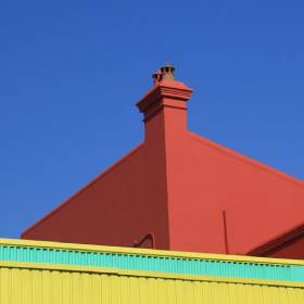 Red chimneys on the top of a red building against a blue sky with the top of a yellow and green building horizontally in front