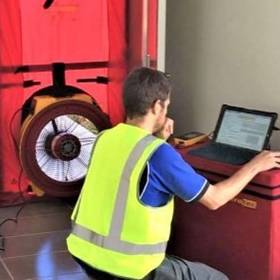 man kneeling and typing on laptop with large fan in background
