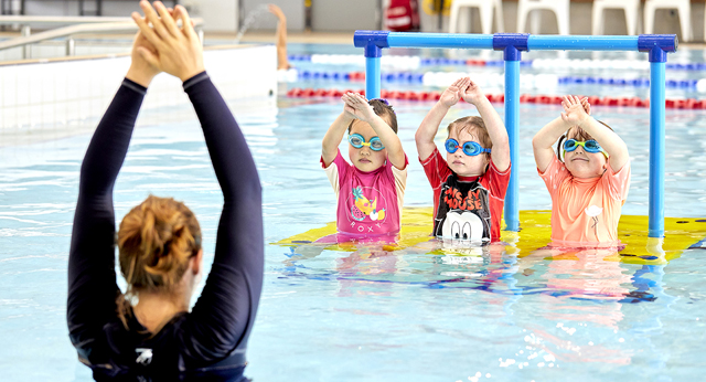 Three children on a swimming platform holding their hands together over their heads in a diving pose copying an instructor