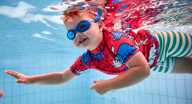 Little boy underwater wearing red rash vest with blue dinosaurs, green and white striped shorts and blue googles