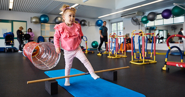 Little girl in a pink top stepping over bar on blue mat in fitness studio with kids exercise equipment set up around the room