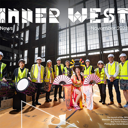 A performer unfurls two decorative fans in front of a group of people wearing high-visibility vests and hard-hats in a tall hall-like ex-industrial space.