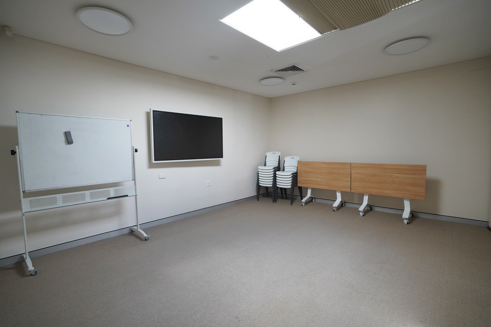 A meeting room with a portable whiteboard, and a flat LCD screen affixed to the wall. A table and stacks of chairs are stored against one wall.
