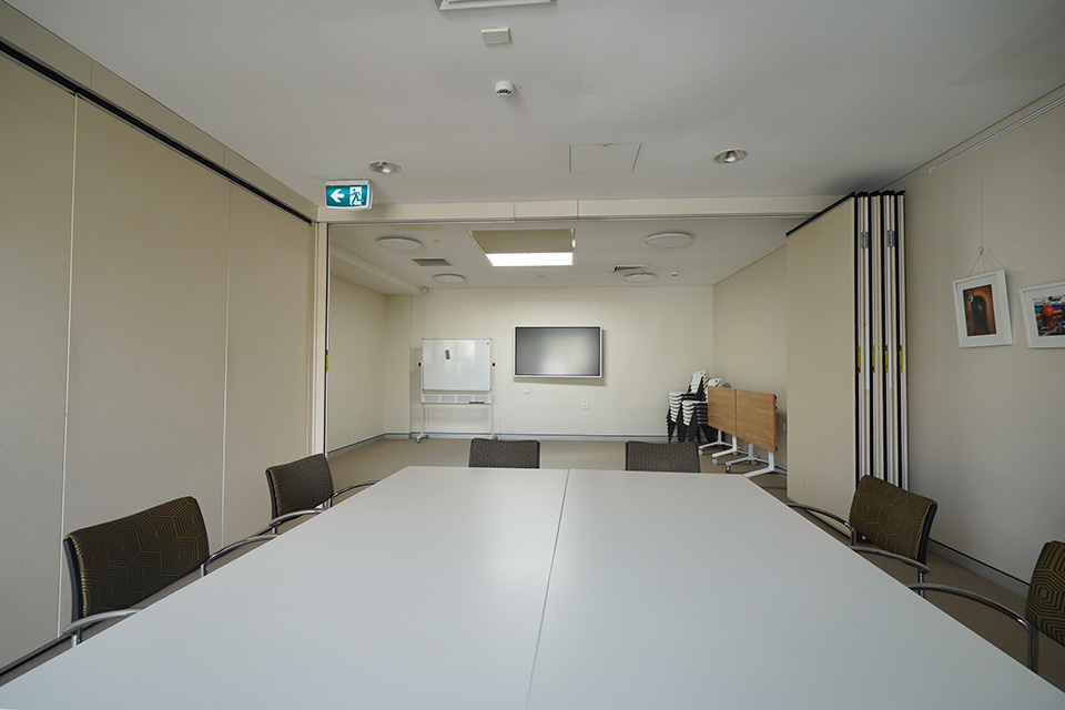 A long meeting room with a whiteboard, a flat LCD screen affixed to the back wall, a table and stacked chairs against one wall, and a meeting setting arranged in the foreground.