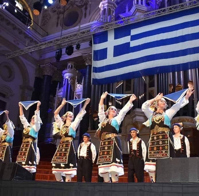 People aligned on stage performing a dance in traditional Greek attire. The Greek national flag is hanging behind the performers.