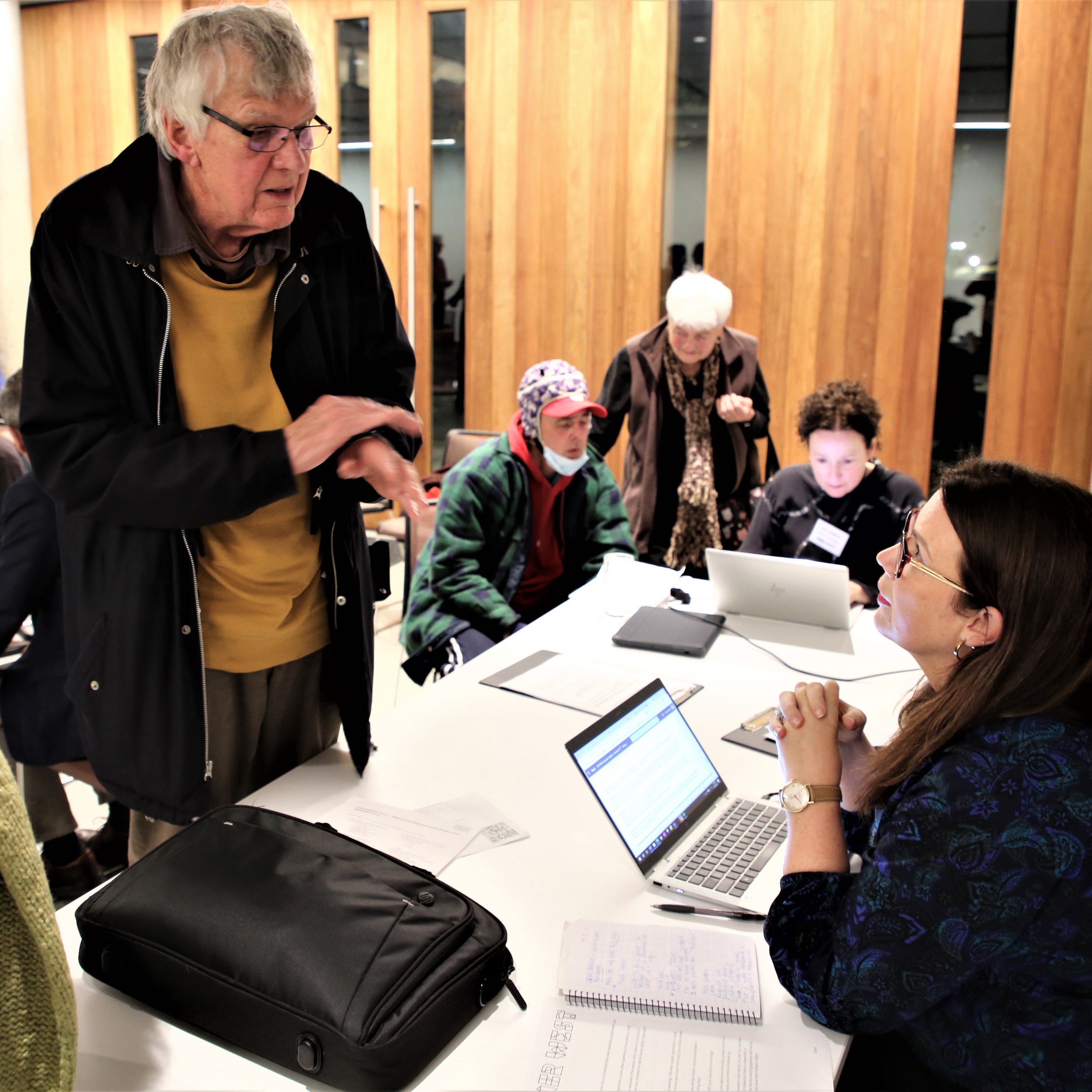 A person sitting at a desk with a laptop talks to a eldery person standing