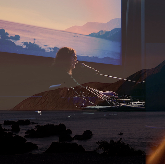 A person performing on electronic musical equipment, faintly interspersed with landscape imagery in the foreground and background.