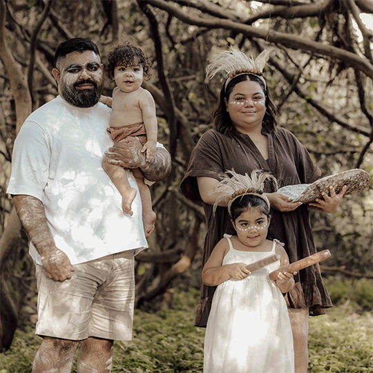 A family of two adults and four children in a bush setting. All four are wearing traditional Aboriginal body paint.