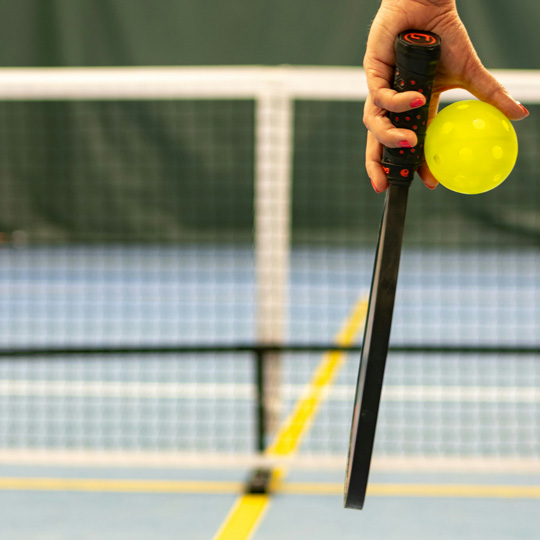 A hand holding a pickleball racket and ball in front of a net