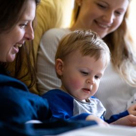 Two adults smiling and reading a book to a child.