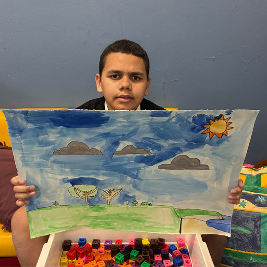 School student holding a watercolour painting of a green hill under a blue sky with a sun in one colour. In front of the painting are some coloured lego blocks