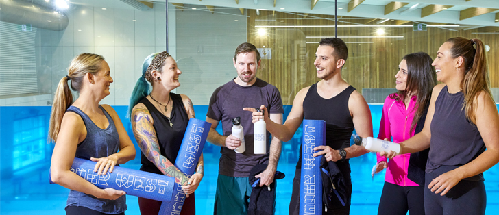 Group of people holding yoga mats and drink bottles in a group fitness studio laughing and smiling at each other
