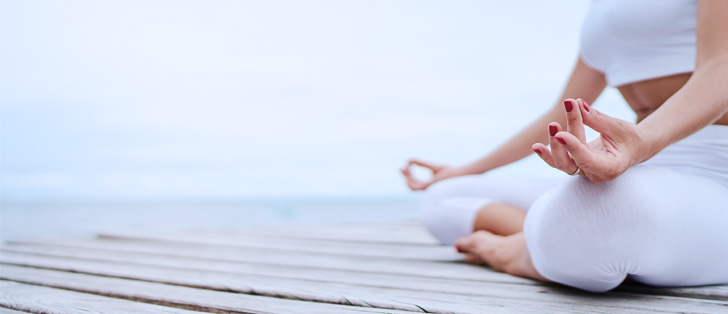 Woman sitting cross legged on wooden deck outdoor with hands on knees in a yoga pose