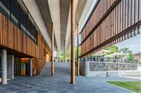 Grand outdoor foyer area with large wooden poles reaching to a white angular ceiling with slatted wooden panels on either side cafe area artwork and green space featured on right