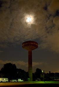 Night scene with bright moon hidden behind clouded sky plus large water tower soaring towards it from green lit grounds