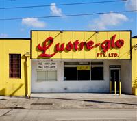 Bright yellow building with the sign Lustre-glo pty ltd in red writing on blue sky with powerlines and road driveway in foreground