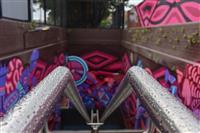 Silver handrails in foreground stretching out and down unseen stairway surrounded by blurred pink and blue graffiti walls