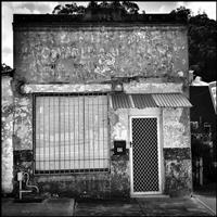 Black and white image of old shop front with peeling paint and faded lettering featuring dark letter box with number 42 and white flyscreen door