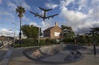 Skate park with two people and graffiti surrounded by trees and an old building on blue clouded sky and airplane soaring closely overhead