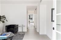 Indoor hallway area leading to kitchen with sitting area in foreground with black and silver armchair black and white rug glass topped coffee table with books and tall white planter with striped foliage plant 