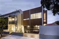 Evening streetfront image taken from slight angle of a contemporary building with wooden slatted frontage small and large rectangular windows and darkened carport below
