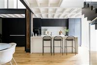 Apartment kitchen interior with warm timber floors black feature wall and cabinetry white island bench with black stools a dining table seen on the left and a dark timber staircase on the right